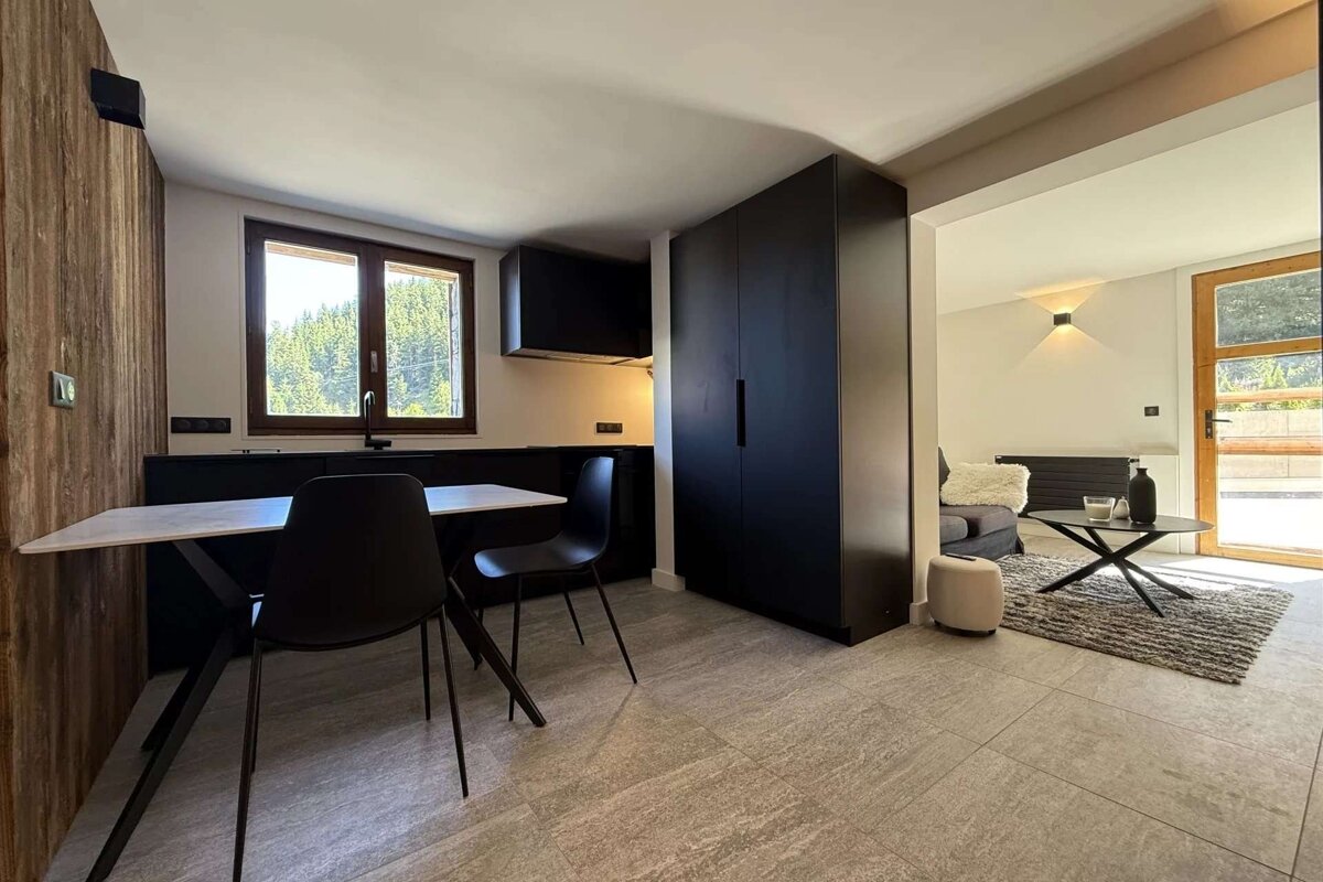A sleek, open-plan apartment featuring a black and white kitchen with forest views, leading into a minimalist living area. Grey tiled floors tie the spaces together.