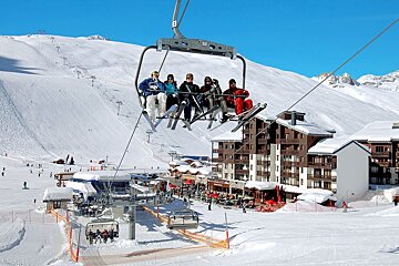 Five skiers ascend on a chairlift over a snowy mountain ski resort with buildings, busy slopes, and other skiers, all under a bright blue sky.