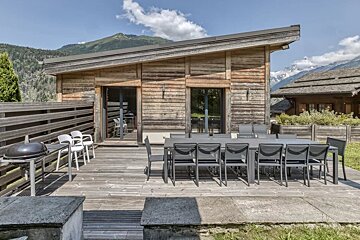 A wooden chalet with a spacious deck featuring a long dining table, a grill, and a mountain backdrop.