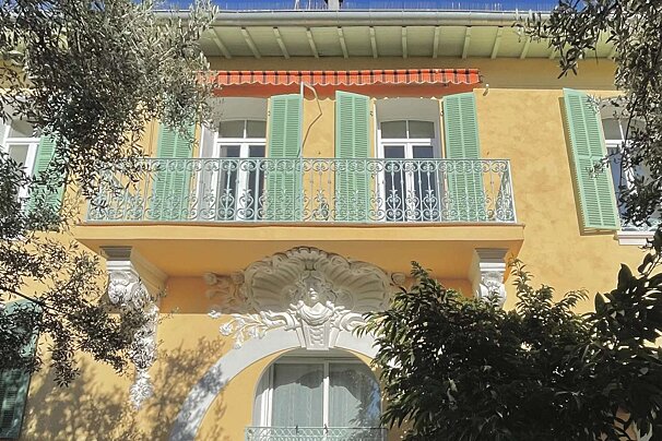 A sunny yellow building with green shutters, an ornate balcony, and decorative white carvings, framed by green foliage.