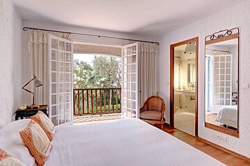 Bright bedroom with a white bed, open French doors to a balcony overlooking trees, and an ensuite bathroom door. Wicker accents & mirror.