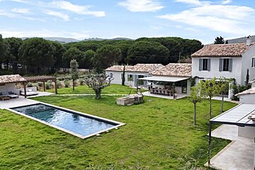 A spacious villa complex with terracotta roofs, a long pool, and manicured lawns, backed by a lush forest under a clear sky.
