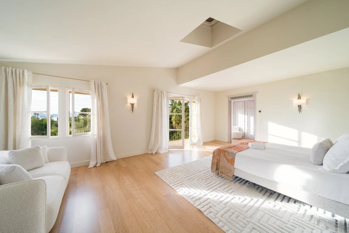 A bright, minimalist bedroom with light wooden floors, a large bed, and a white sofa. It features windows overlooking greenery and the ocean, plus a skylight.