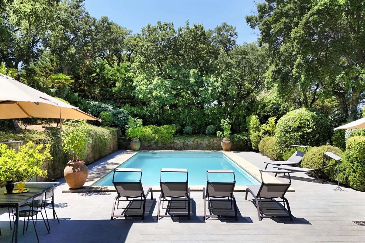 Sunny outdoor pool area with lounge chairs, dining set, and lush green trees and bushes, creating a serene, relaxing retreat.