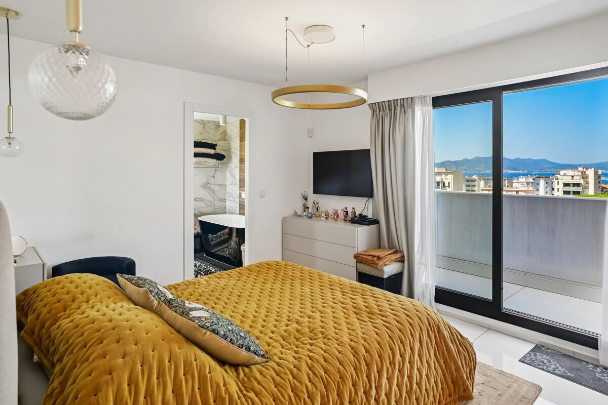 Modern bedroom with a mustard-yellow quilted bed, a sliding glass door to a balcony with sea views, and a doorway to a chic bathroom.
