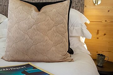 A bed styled with a textured beige throw pillow, white linens, a Robb Report magazine, and a coffee mug on a bedside table.