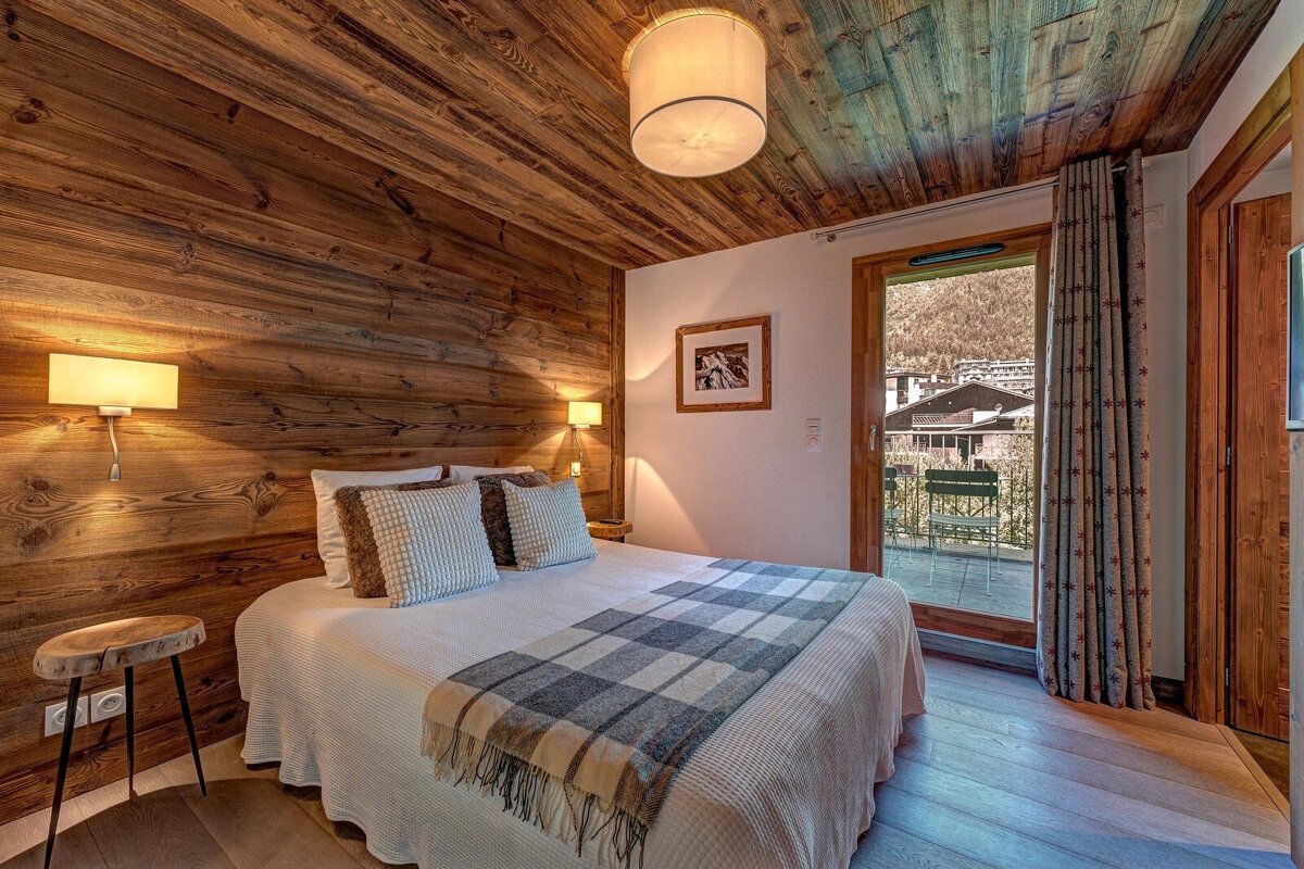 Cozy rustic bedroom with wood walls and ceiling, a comfortable double bed, and glass doors opening onto a scenic balcony.