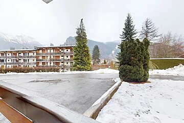 A winter scene in a mountain town, showing apartment buildings with balconies, evergreen trees, snow-covered ground, and distant mountains under a grey sky.