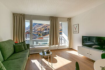 Bright living room with a green sofa, TV, and large windows revealing a mountain village nestled among golden autumn trees.