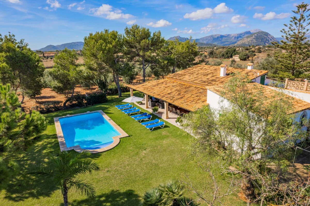 Aerial view of a sunny villa with a pool, green lawn, sun loungers, and surrounding trees, set against a backdrop of distant mountains.