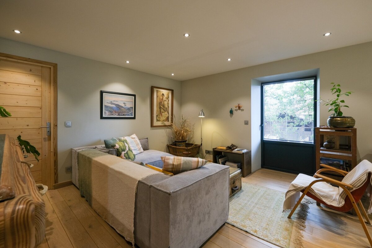 A cozy living room with a light grey L-shaped sectional sofa, wooden floor, and a door. Artwork adorns the walls, and a window with blinds provides light.