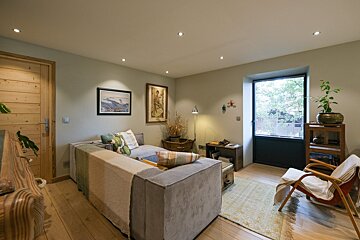 A cozy living room with a light grey L-shaped sectional sofa, wooden floor, and a door. Artwork adorns the walls, and a window with blinds provides light.