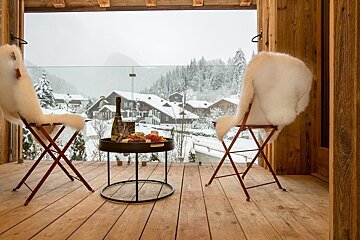 Cozy wooden balcony with sheepskin-covered chairs and wine overlooking a snowy alpine village.
