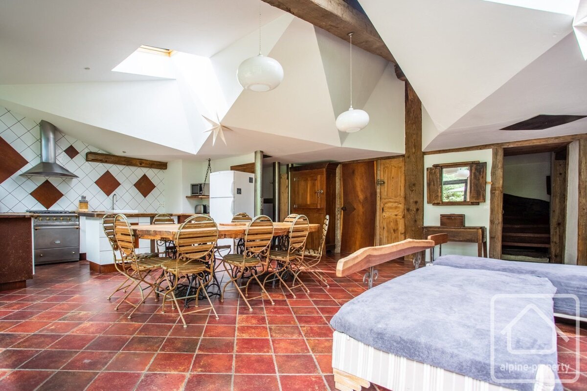 An open-plan rustic room with terracotta tiles, wood beams, a modern kitchen, large dining table, and daybeds under bright skylights.