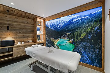 A rustic-modern spa room features a massage table, wooden walls, and a large mural of snowy mountains overlooking a vibrant turquoise lake.