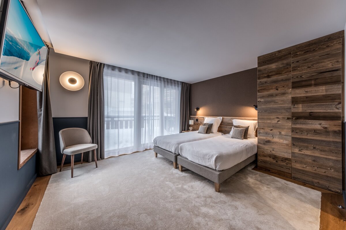 Cozy hotel bedroom with two twin beds, rustic wood paneling, an armchair, and a TV, featuring large windows and soft, warm lighting.