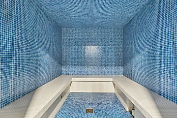 An empty blue mosaic-tiled steam room or hammam with white tiled benches along the walls and a central floor drain.