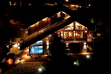 A luxurious, multi-level chalet glowing brightly at night with an indoor pool visible through large windows and snow-covered ground.