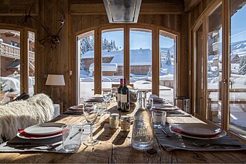 Rustic dining room in a cozy chalet, with a set wooden table and large windows revealing a sunlit, snow-covered mountain village scene.