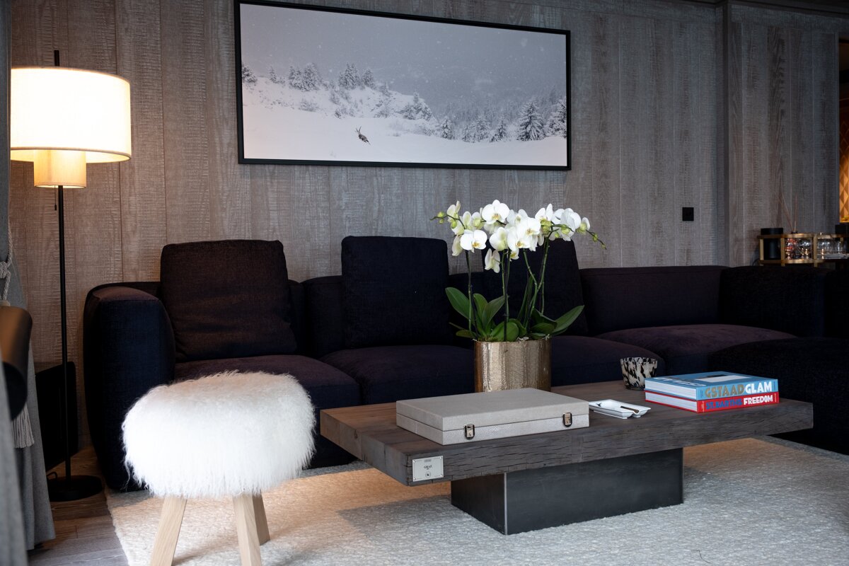 A modern living room featuring a dark sofa, wooden coffee table with white orchids, fluffy stool, and a large winter landscape artwork on a paneled wall.