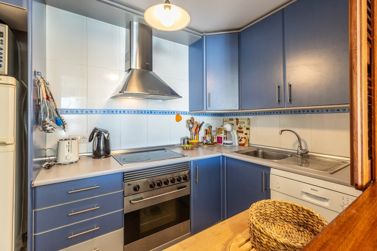 A small, functional kitchen featuring blue cabinets, white tiled walls, a stainless steel range hood, stove, sink, and dishwasher.