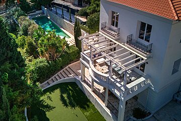 Aerial view of a luxurious white house with a red roof, multiple terraces under pergolas, a swimming pool, a putting green, and lush gardens.
