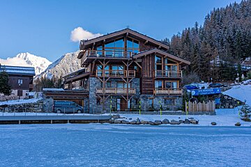 A luxurious wooden and stone chalet with balconies overlooks a frozen surface, likely a lake or rink, with snow-capped mountains and pine forests in the background.