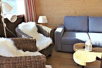 A cozy living room with wooden walls, wicker chairs with fur throws, a gray sofa, and rustic wood tables, illuminated by sunlight.