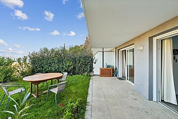 An outdoor patio of a building overlooks a grassy yard with a wooden dining table and chairs, set against a blue sky.