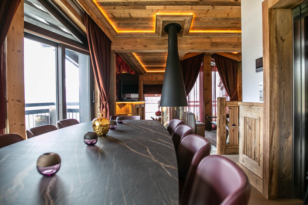 A luxurious, rustic-modern dining room features a long table, maroon chairs, a stylish hanging fireplace, and warm wooden decor with accent lighting.
