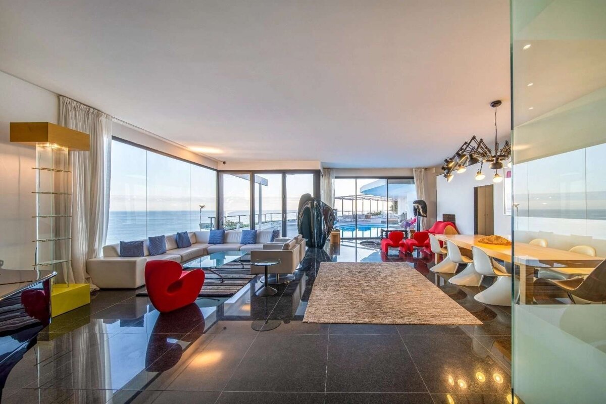 A luxurious, modern living room with floor-to-ceiling windows offers stunning ocean views and a pool. It features stylish furniture and a shiny dark floor.