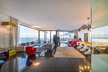 A luxurious, modern living room with floor-to-ceiling windows offers stunning ocean views and a pool. It features stylish furniture and a shiny dark floor.