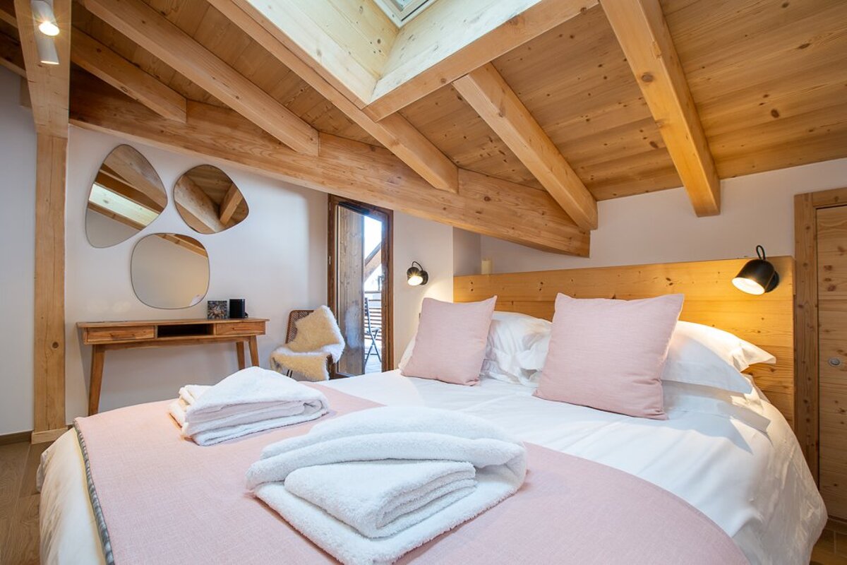 Inviting bedroom with a wood-beamed ceiling, large bed with pink and white linens, fresh towels, a wooden desk, and a balcony door.