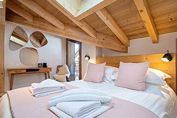 Cozy chalet bedroom with exposed wooden beams, a bed with pink pillows and white towels, a vanity desk, and organic-shaped mirrors.