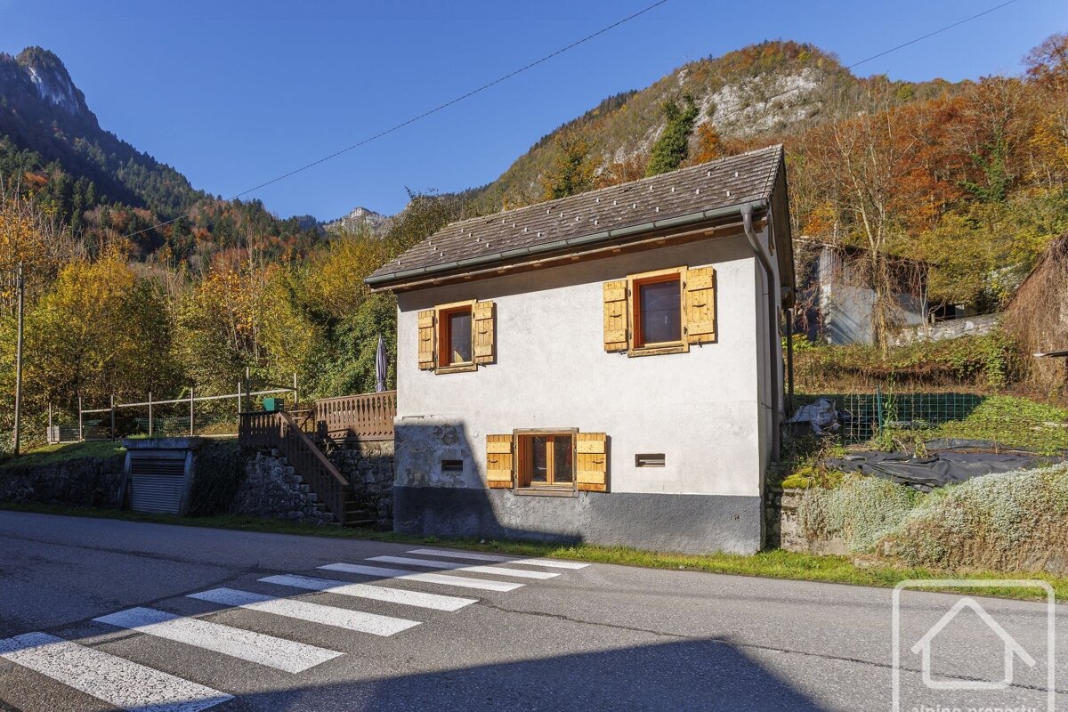 A quaint house with wooden shutters sits by a road with a crosswalk, framed by colorful autumn mountains under a clear blue sky.