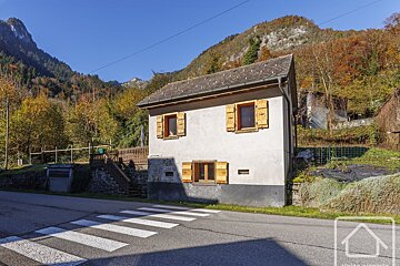 A quaint house with wooden shutters sits by a road with a crosswalk, framed by colorful autumn mountains under a clear blue sky.