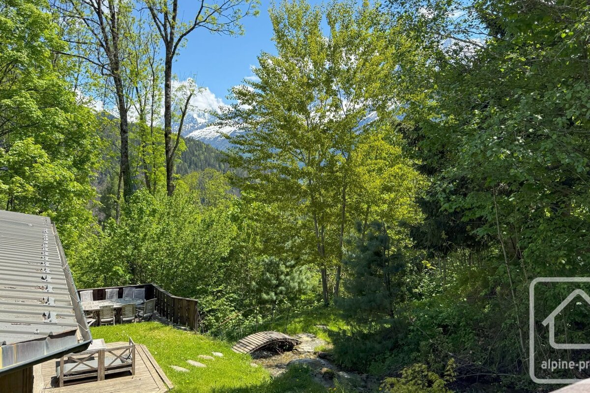Lush green backyard with a deck, patio furniture, a small bridge, and tall trees, with snow-capped mountains in the distance.