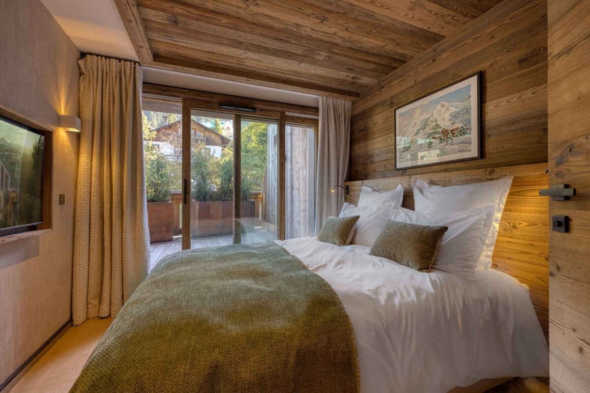 A rustic wood-paneled bedroom with a large bed, white linens, a green throw, and sliding doors opening to a balcony with greenery.