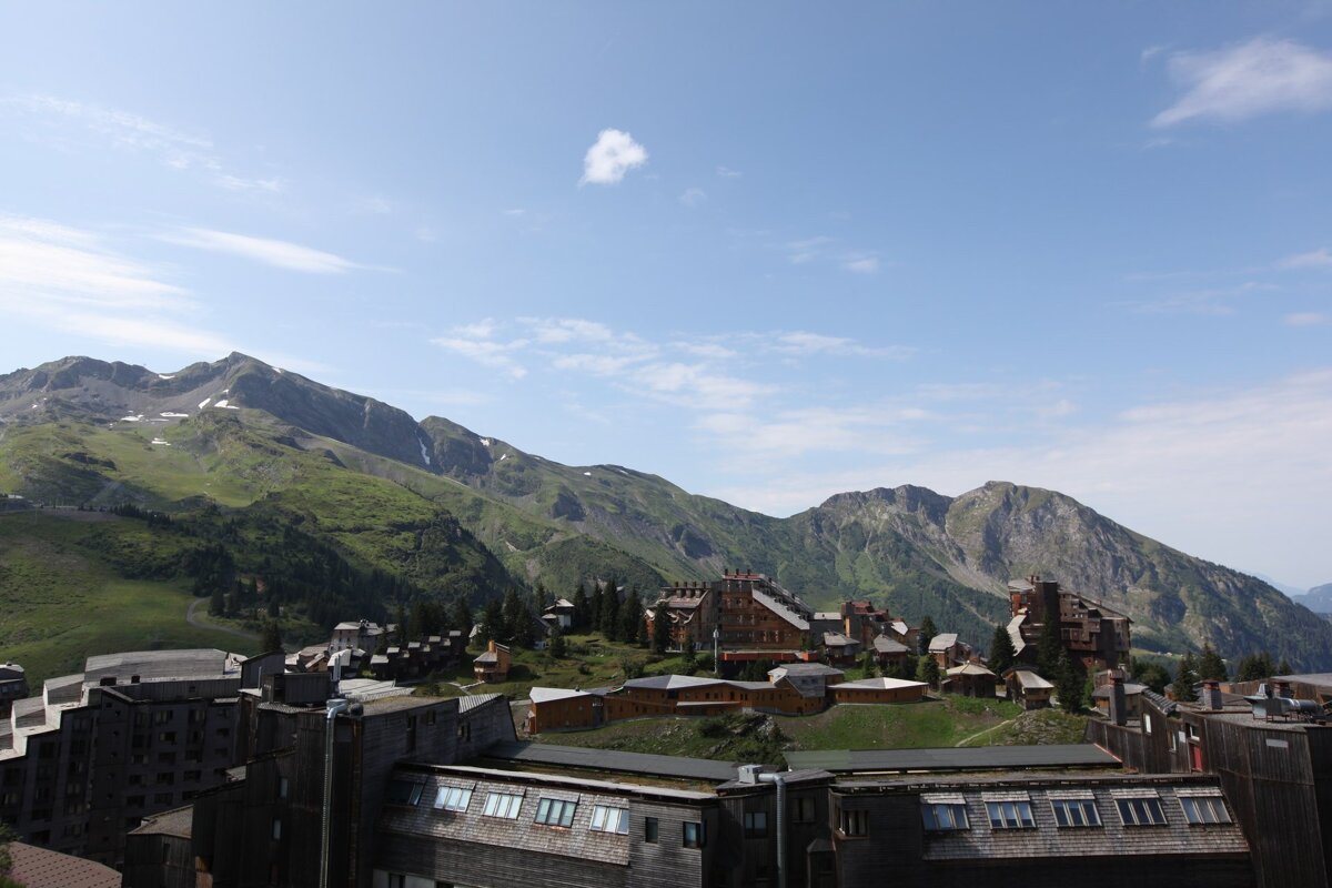 A mountain village with multi-story buildings is nestled among lush green and rocky peaks with patches of snow under a bright blue sky.