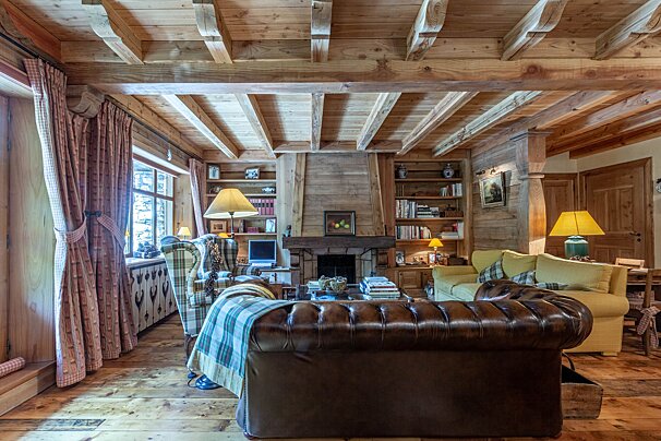 A cozy, rustic living room in a wood-paneled chalet features a stone fireplace, leather and plaid sofas, bookshelves, and large windows.