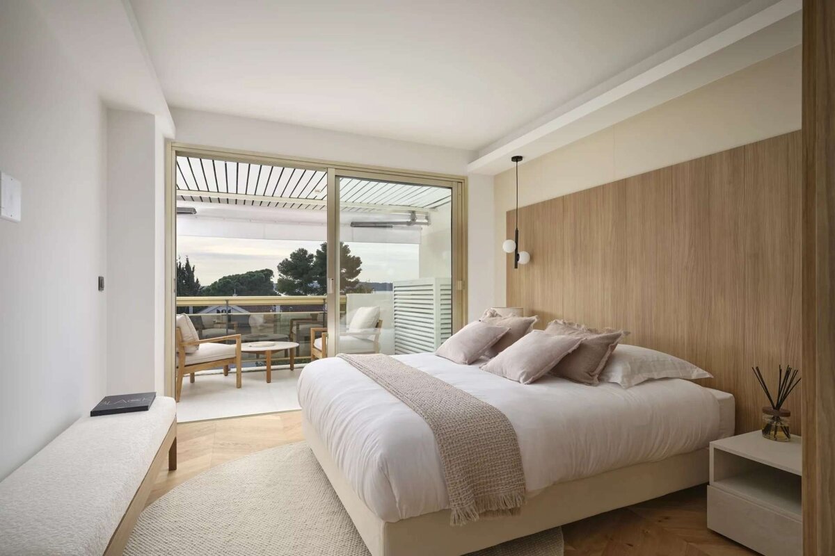 A serene, modern bedroom with a large bed, light wood features, and a glass door opening to a private balcony with outdoor furniture and a view.