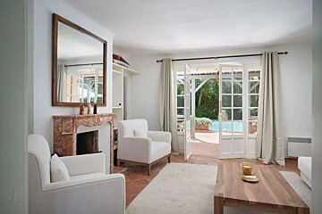 A bright living room with white chairs, a fireplace, and French doors opening to a patio with a swimming pool and lush greenery.