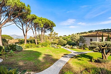 Lush garden with pine trees, paved path, traditional house, and distant sea view under a clear blue sky.