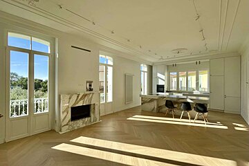 A sunlit, modern living space with white walls, herringbone wood floors, a marble fireplace, and a sleek kitchen island. Large windows offer green views.