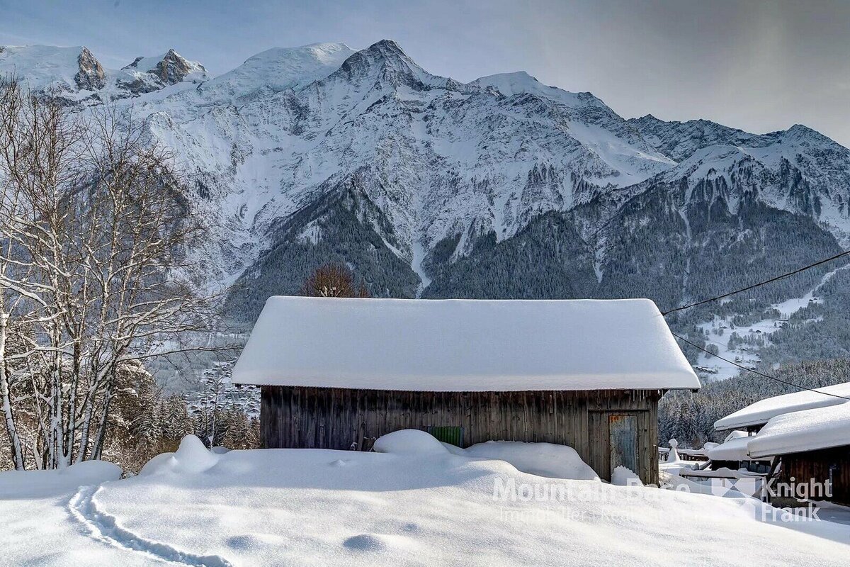 A rustic wooden cabin blanketed in snow, with footprints in the foreground. Towering snow-capped mountains and alpine forests create a majestic winter scene.