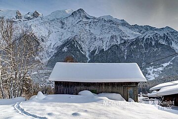 A rustic wooden cabin blanketed in snow, with footprints in the foreground. Towering snow-capped mountains and alpine forests create a majestic winter scene.