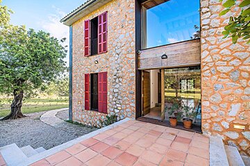 A rustic stone house with red shutters, large glass windows, and a tiled patio. A tree and greenery are visible on the left.