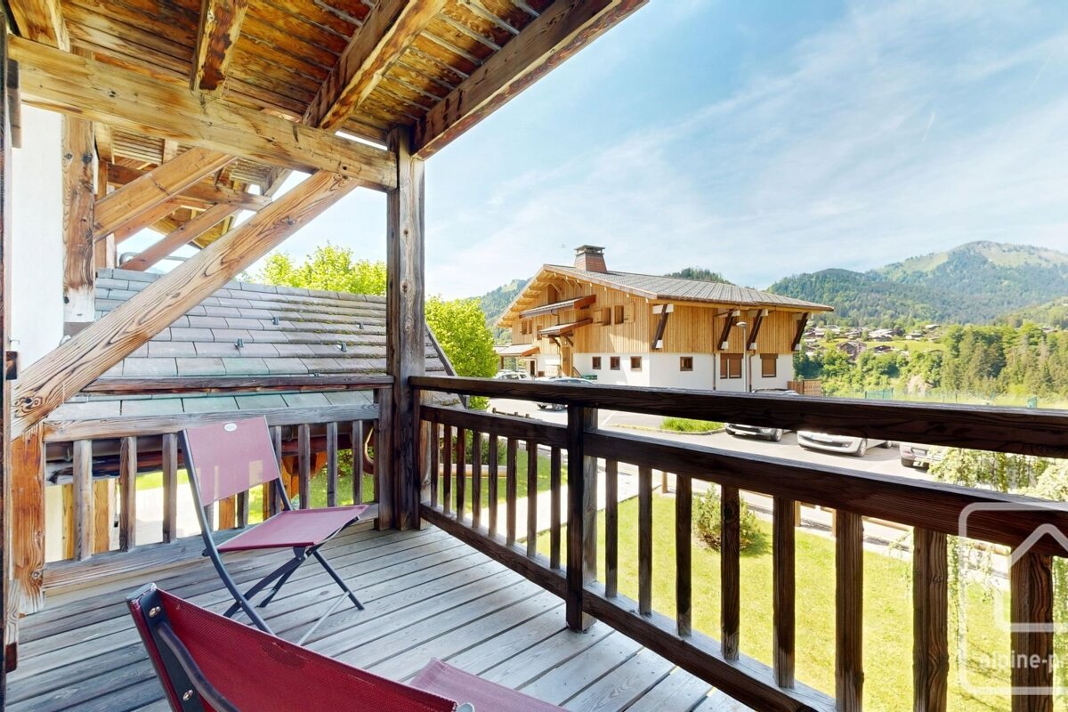 Wooden balcony with chairs overlooking a mountain village chalet and green hills under a bright blue sky.