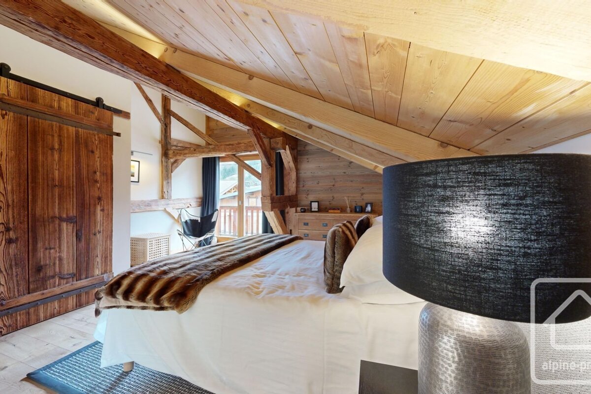 Cozy chalet bedroom with wood beams, a large bed with a fur throw, a sliding barn door, and a balcony view.