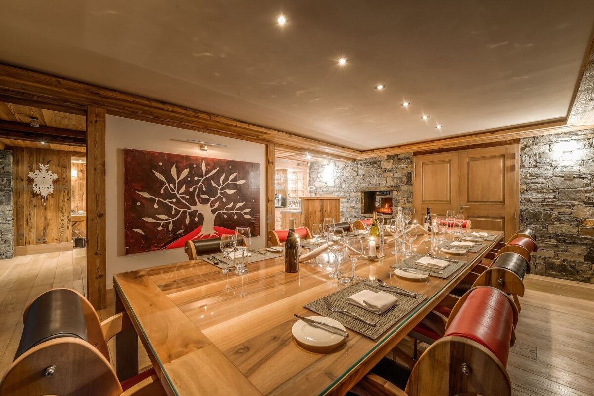 Elegant chalet dining room with a long wooden table, unique barrel-back chairs, stone walls, a cozy fireplace, and a tree artwork.